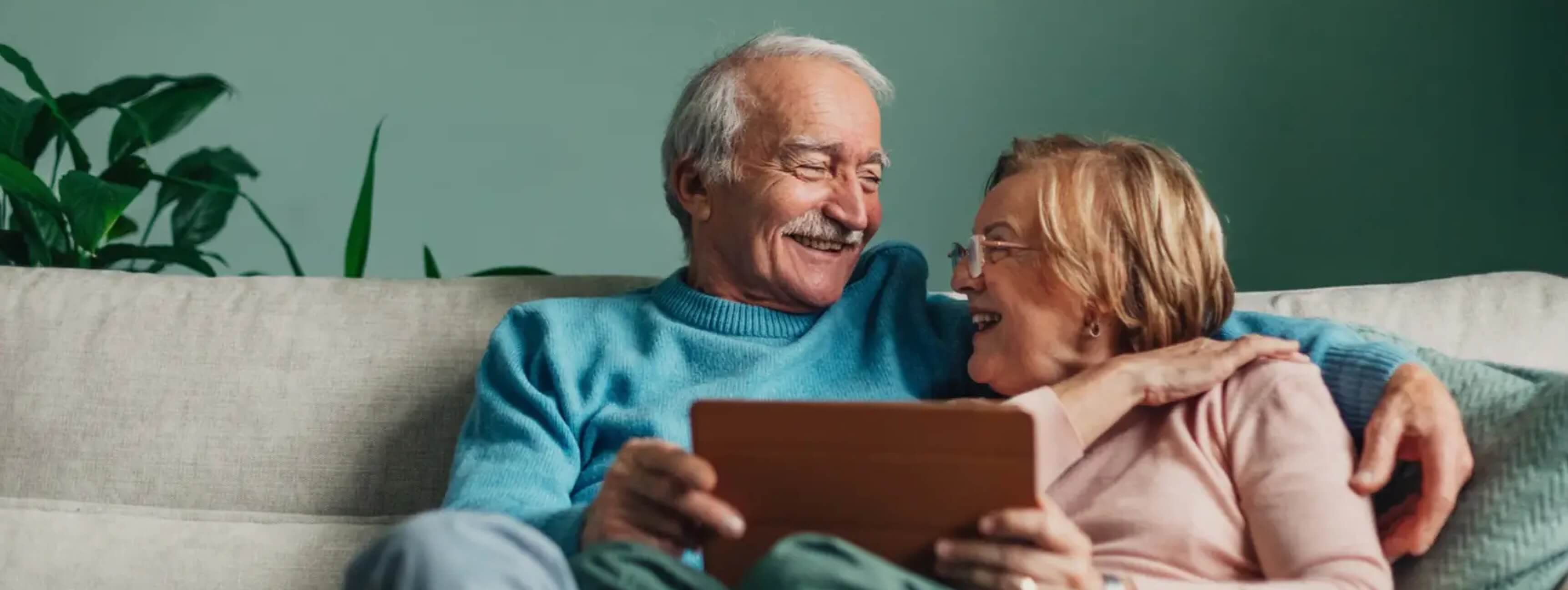 An elderly couple sitting on a sofa smiling at each other whilst holding a digital tablet